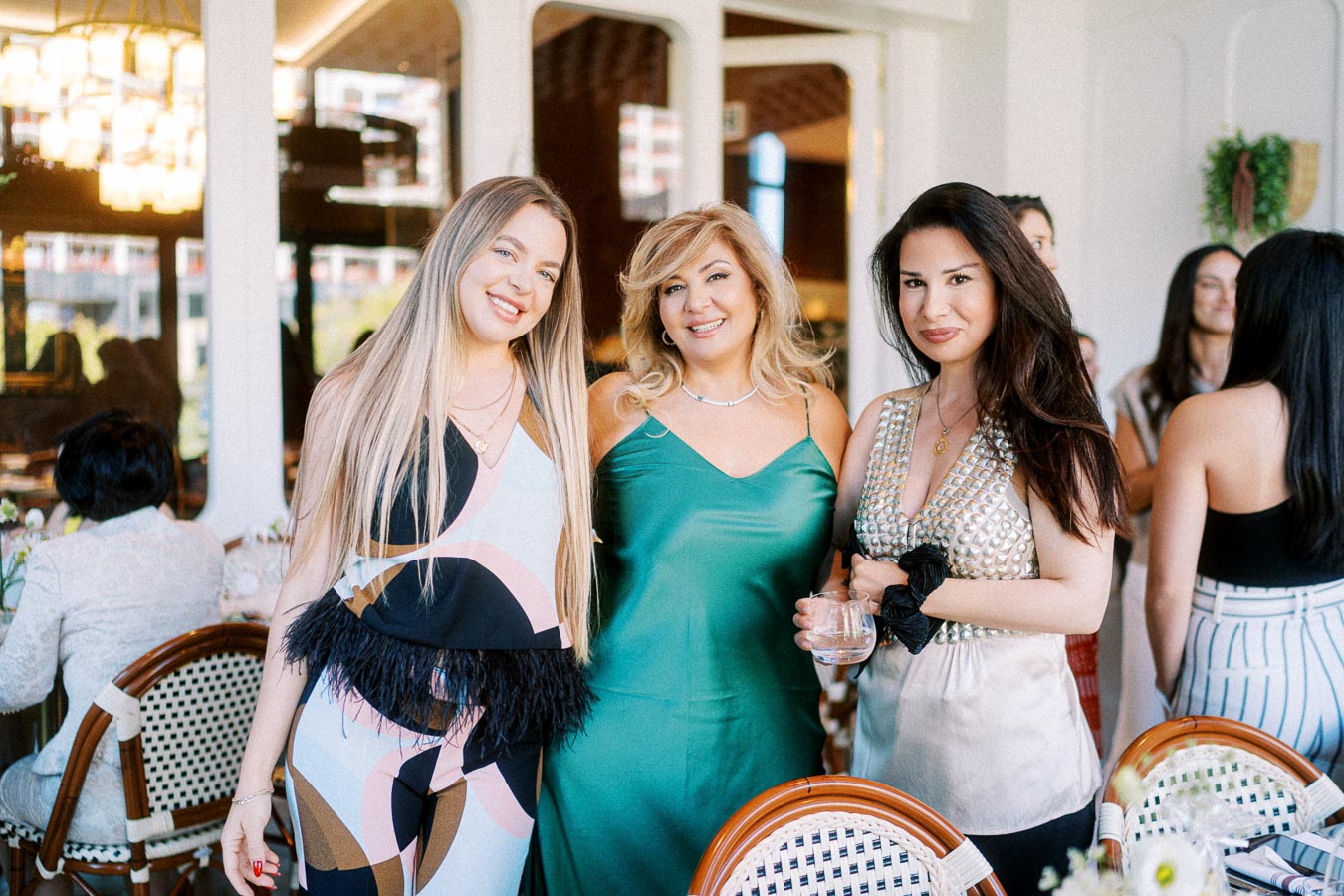 Three women smiling and posing together indoors at a stylish brunch event, dressed in colorful and elegant outfits.