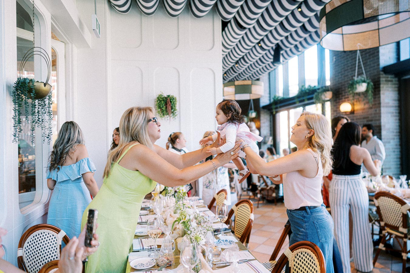 A lively family gathering in a stylish restaurant. Adults in colorful attire are playfully interacting with a cheerful baby, surrounded by elegantly decorated tables and hanging plants, creating a warm and inviting atmosphere.
