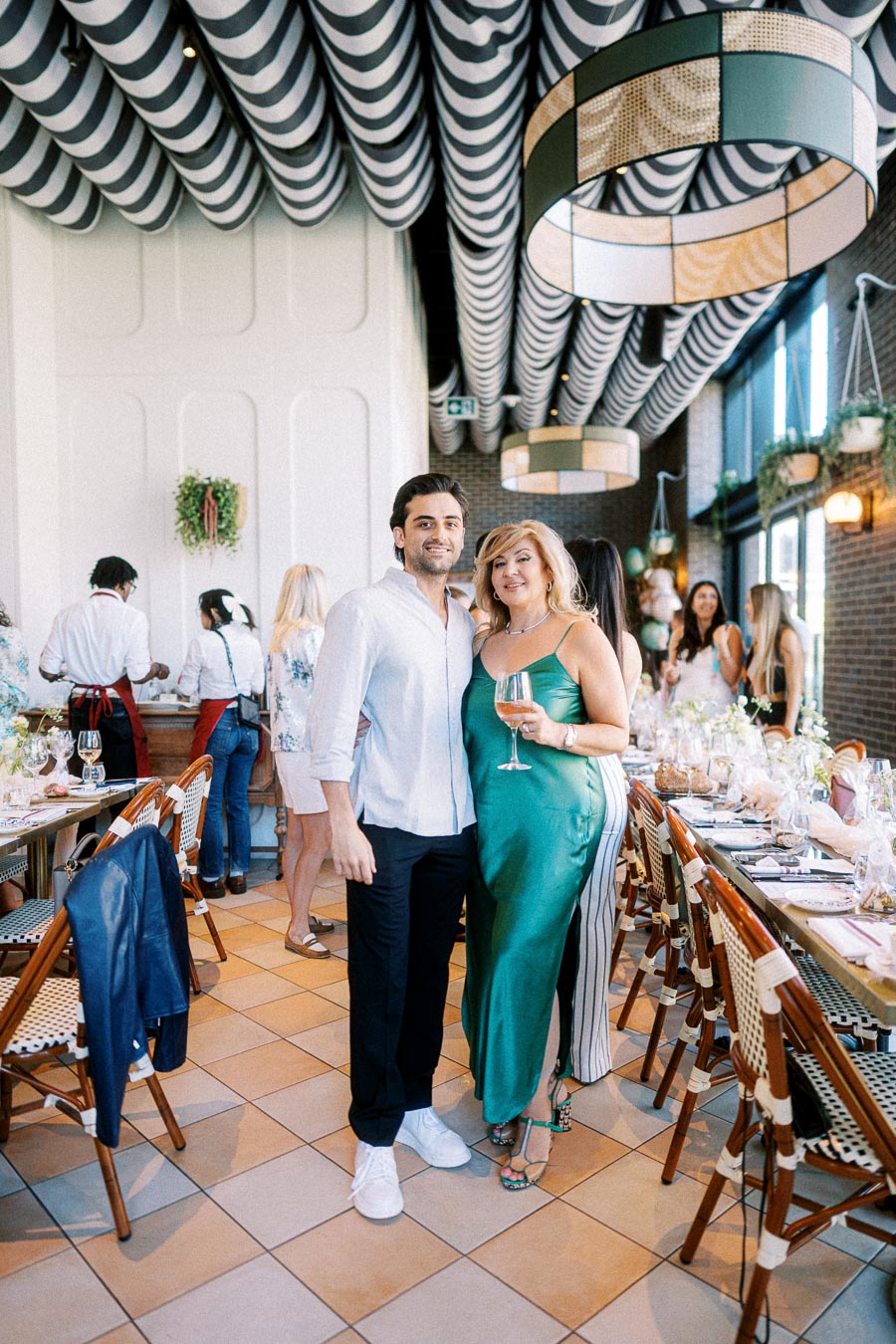 A couple posing together at a stylish indoor event, with elegantly set tables and a modern decor backdrop, featuring striped ceilings and hanging lights.