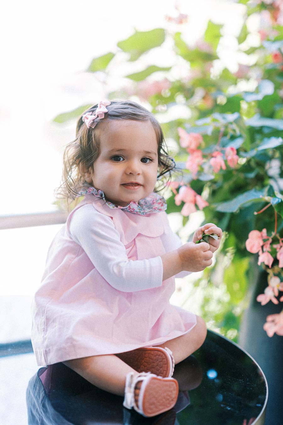 A toddler in a light pink dress and matching hair bow sits happily on a reflective surface, surrounded by vibrant green leaves and pink flowers, in a sunny outdoor setting.
