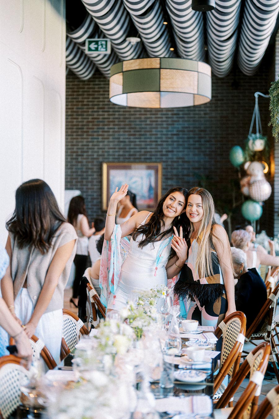 Two women smiling and waving at a well-decorated brunch event, with a table set with elegant white flowers and tableware, against a stylish interior with patterned ceiling and hanging decor.