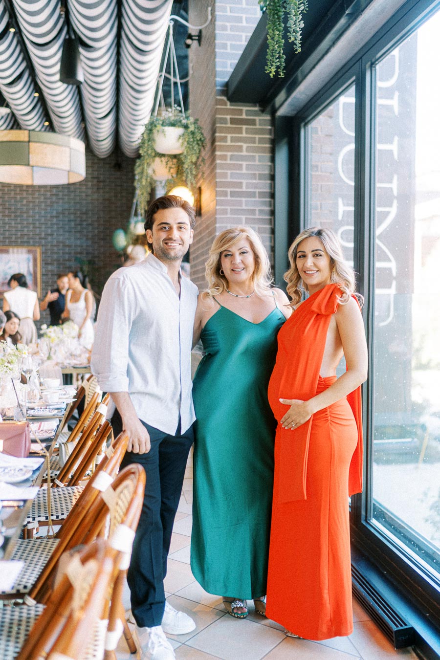 Three people smiling at a stylish indoor gathering, with a well-decorated table set for a meal. The person on the right is wearing a bright orange dress and cradling their baby bump, conveying a celebratory atmosphere.