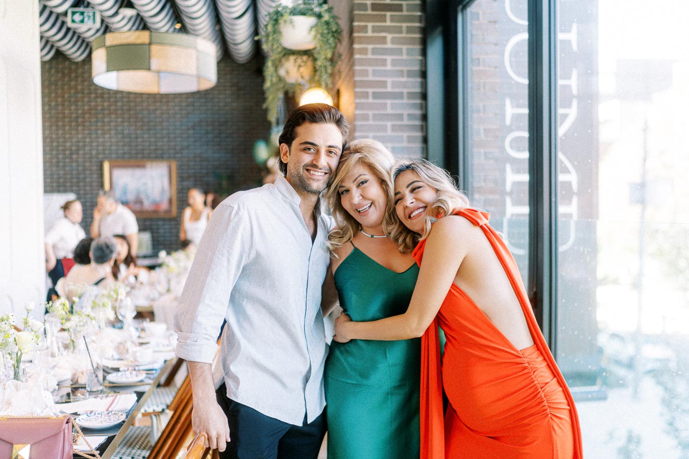 Three people smiling and embracing in a bright restaurant setting, with elegantly set tables and modern decor in the background.