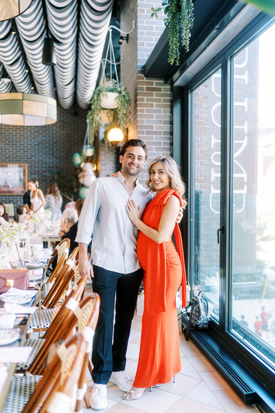 A couple posing happily at a beautifully decorated indoor restaurant, featuring elegant wooden chairs, a brick wall, and hanging plants. The woman is wearing a vibrant red dress, creating a striking contrast with the setting. The large window offers ample natural light, contributing to the cozy and inviting atmosphere.