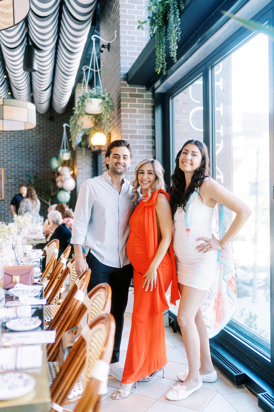 Three people smiling at a stylish indoor event; a pregnant woman in a red dress stands between a man in a light blue shirt and a woman in a white dress, with a beautifully set table in the foreground.