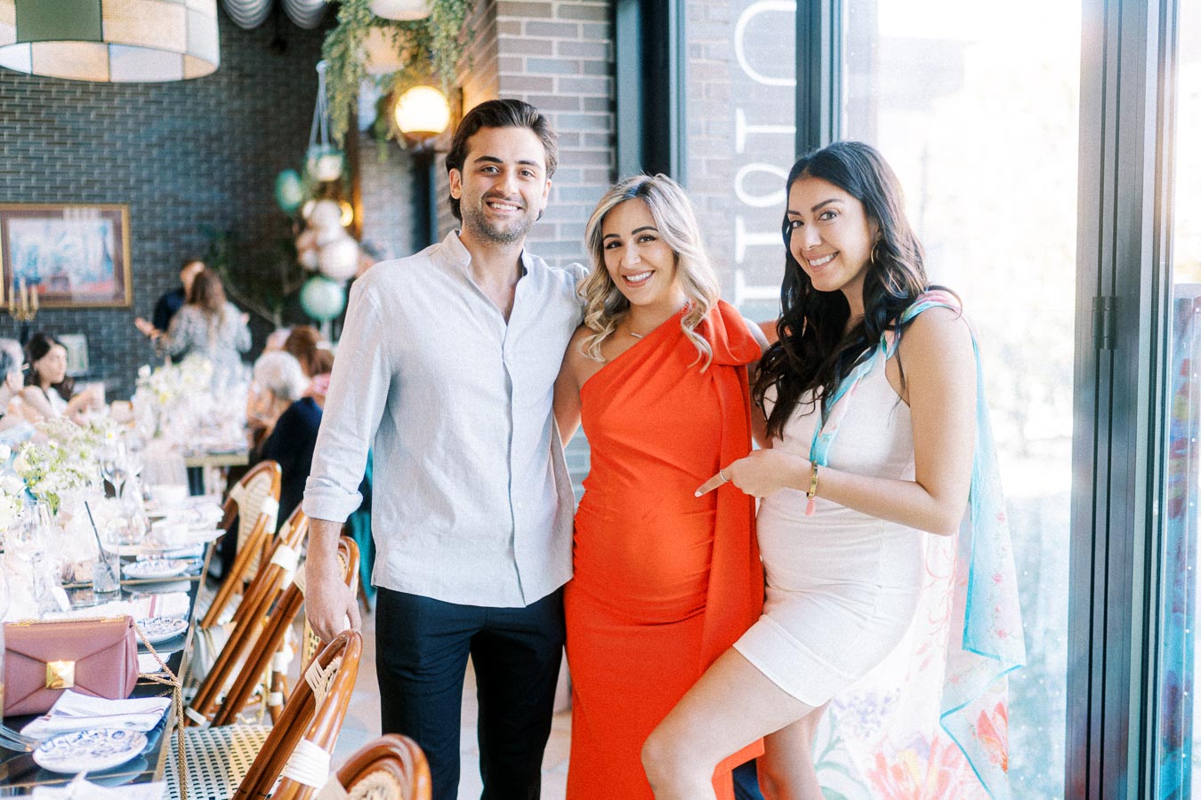 Group of three friends smiling at a stylish indoor restaurant event, with a decorated table and pendant lights in the background.