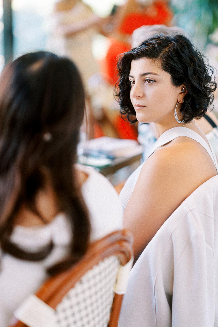 Woman with short curly hair and hoop earrings looking attentively at another person in a social setting, wearing a white top.
