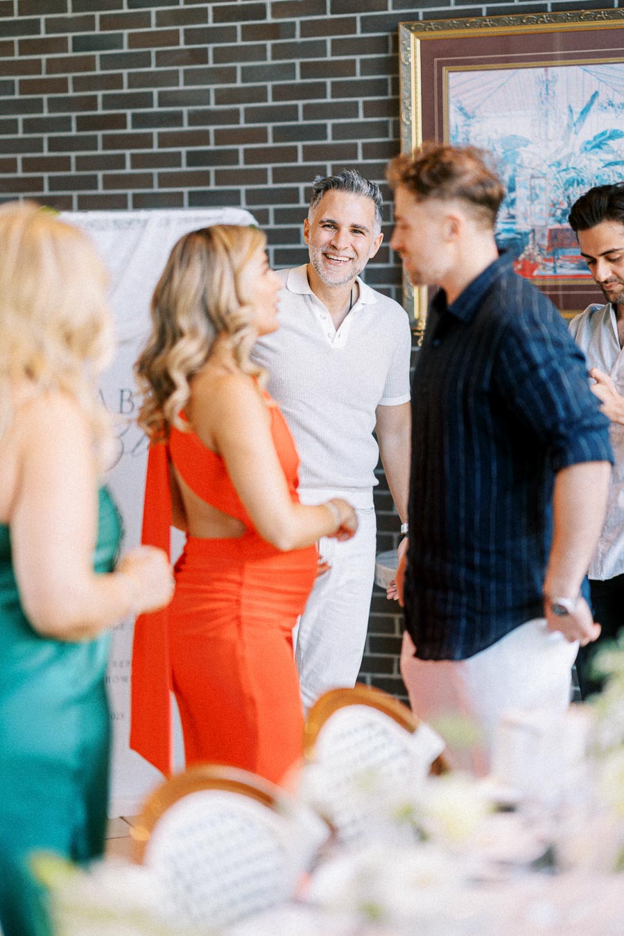 A group of people engaging in a lively conversation at a social gathering, with a woman in a red dress and others dressed in casual attire, standing in an indoor setting with a brick wall backdrop and art pieces.