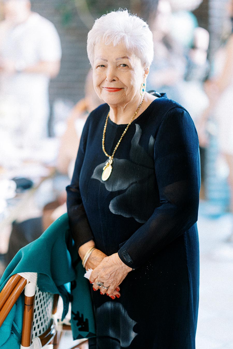 Elderly woman in elegant navy dress with white hair and jewelry, smiling in a warmly lit indoor setting.