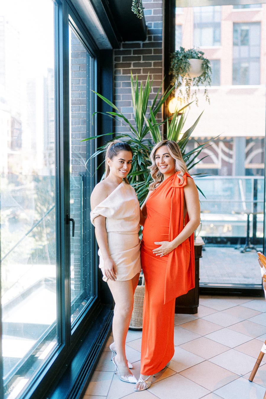 Two women standing indoors, one in a beige dress and one pregnant in an orange dress, smiling and positioned by a large window with a potted plant in the background.