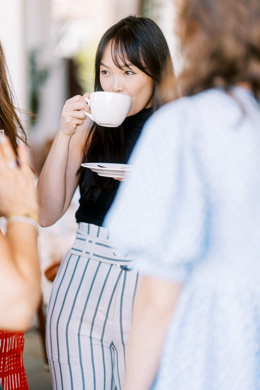 A woman sipping coffee at a social gathering, dressed in a sleeveless black top and high-waisted striped pants, with blurred people in the background enhancing the casual and lively atmosphere.