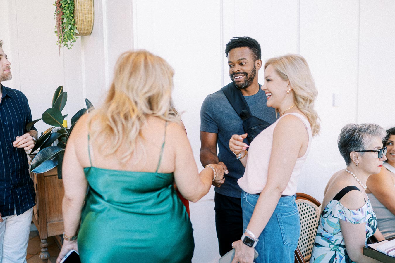 Group of people interacting and smiling at a casual indoor gathering, with a focus on two women shaking hands with a man, surrounded by others seated and standing, green plants in the background.