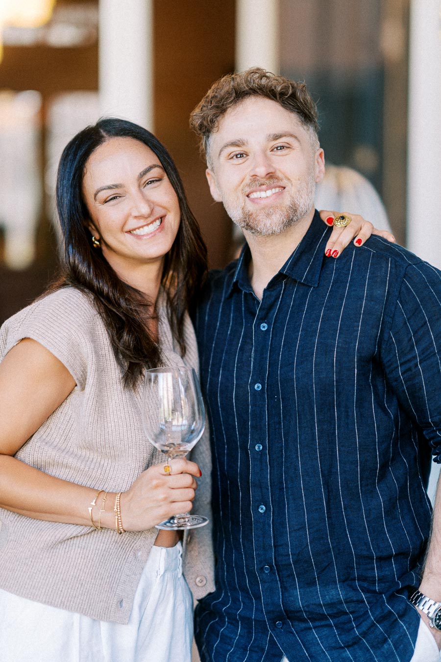 Smiling couple enjoying a social gathering indoors, with the woman holding a wine glass and wearing a beige sweater, and the man in a navy blue pinstripe shirt.