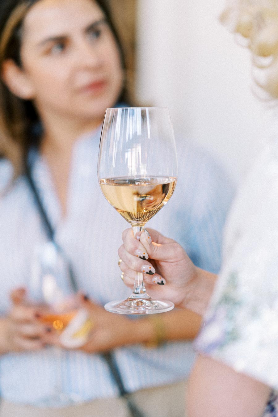 Close-up of a person holding a glass of white wine, with a blurred background showing a social gathering atmosphere.