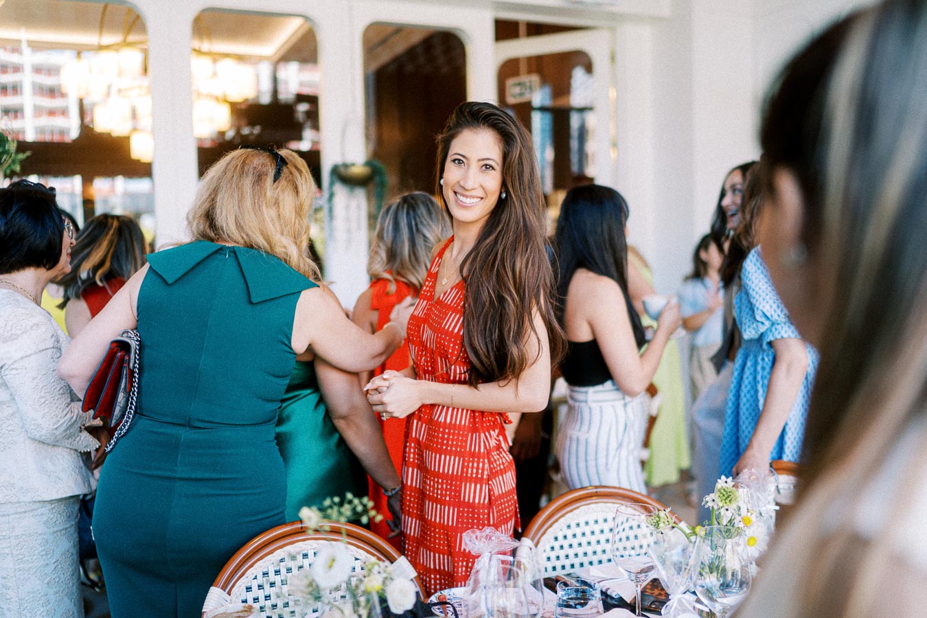 A group of women socializing at an elegant indoor event, featuring a smiling woman in a red dress in the center, surrounded by guests in colorful attire, with floral arrangements and table settings visible in the foreground.