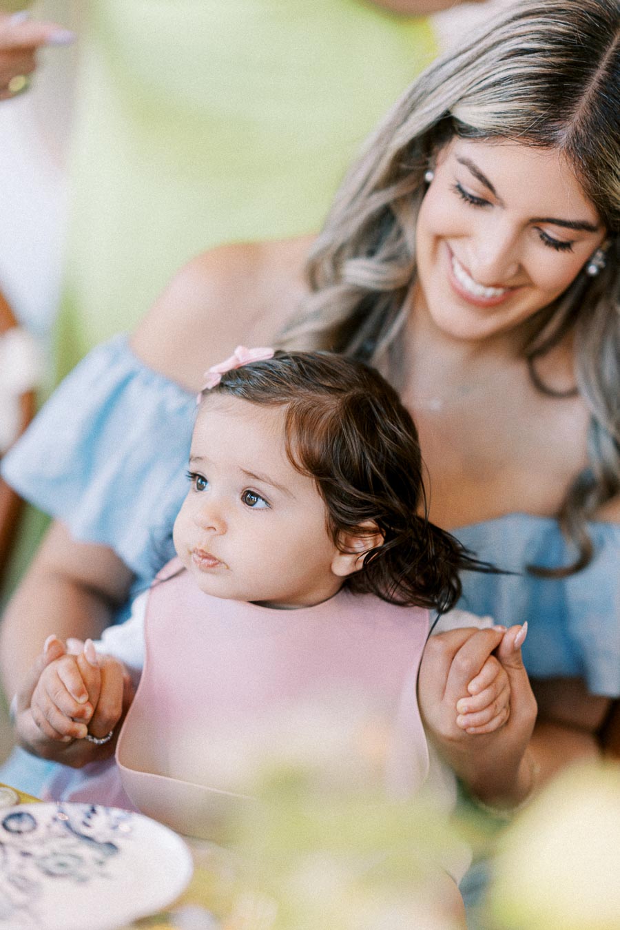 Smiling woman holding a baby wearing a pink bib at a party, captured in a candid, joyful moment.