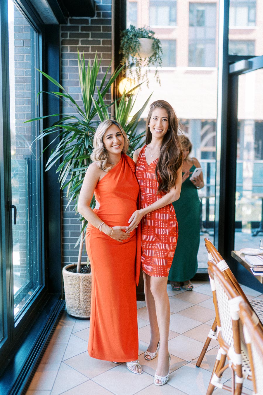 Two women smiling at a baby shower, one pregnant in an elegant orange dress and the other in a stylish red dress, standing in a sunlit room with a green plant in the background.