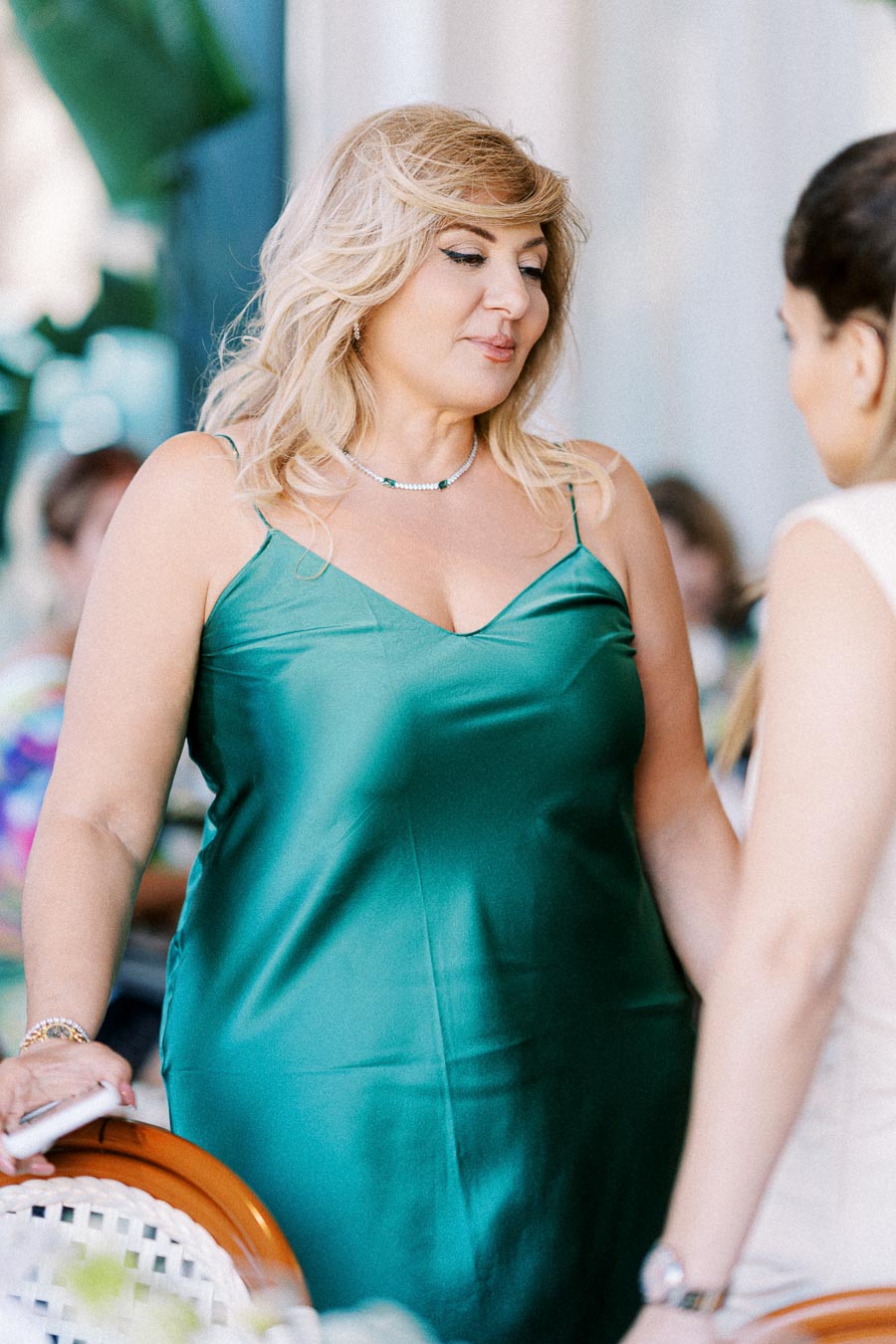 A woman in a green satin dress engages in conversation at a social event, surrounded by blurred greenery and soft lighting in the background.