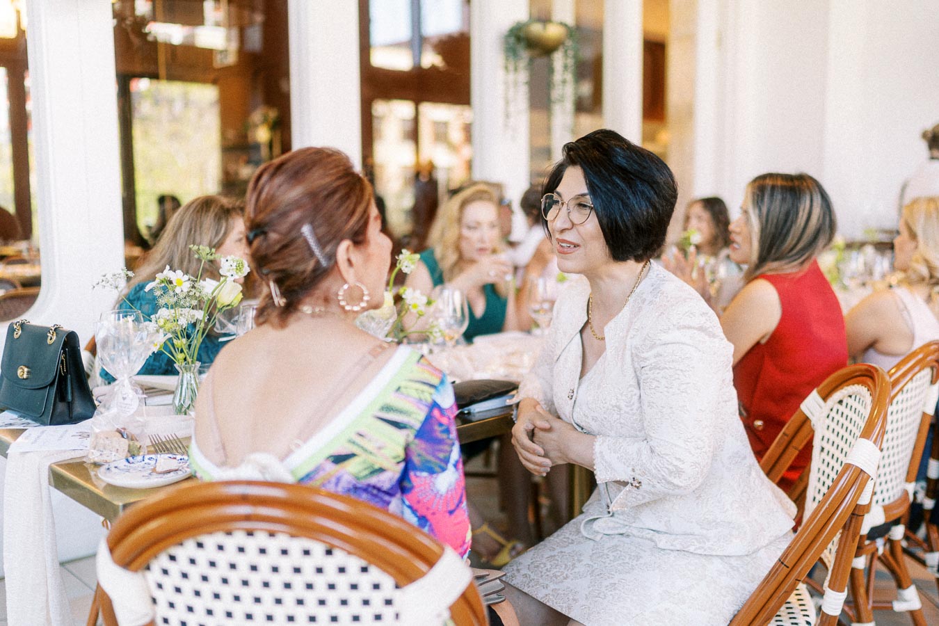 Group of women engaged in conversation at a stylish indoor gathering, seated around a table with elegant tableware and floral decor.