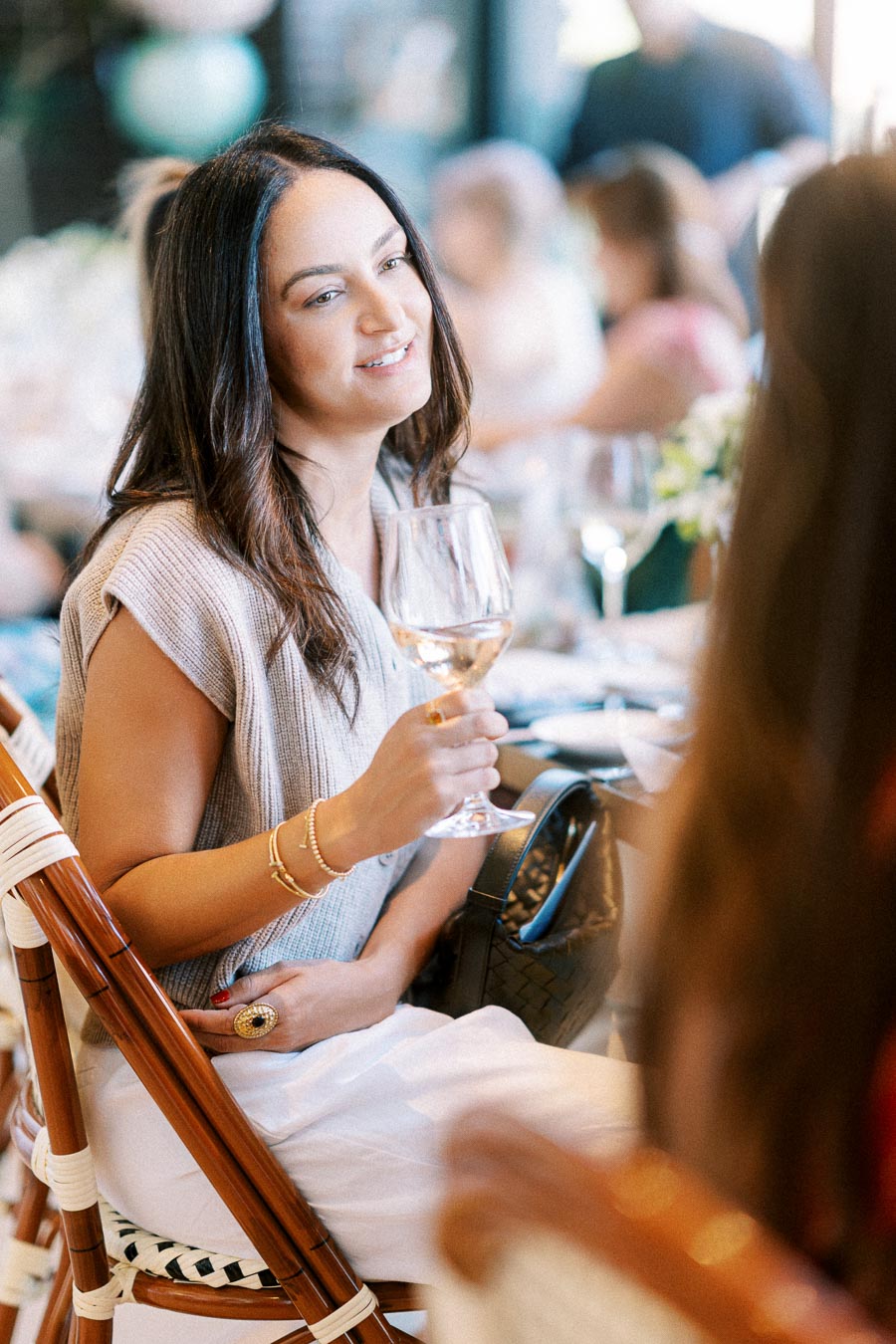 A woman enjoying a social gathering, seated with a glass of white wine in hand, surrounded by a softly blurred background of people, highlighting a relaxed and elegant atmosphere.