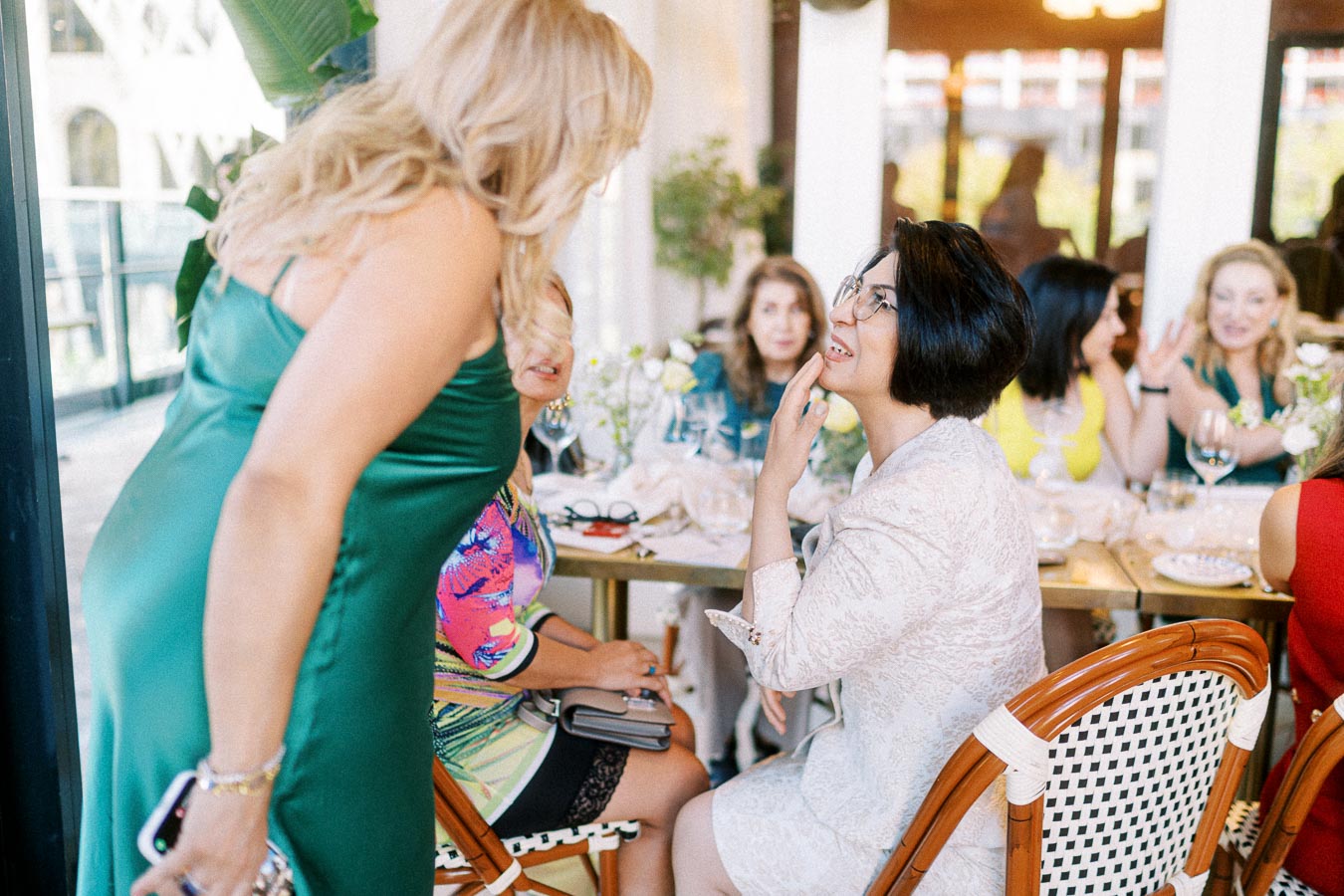 A group of women engaging in conversation at a stylish restaurant gathering, with elegant decor and natural light filtering through large windows.