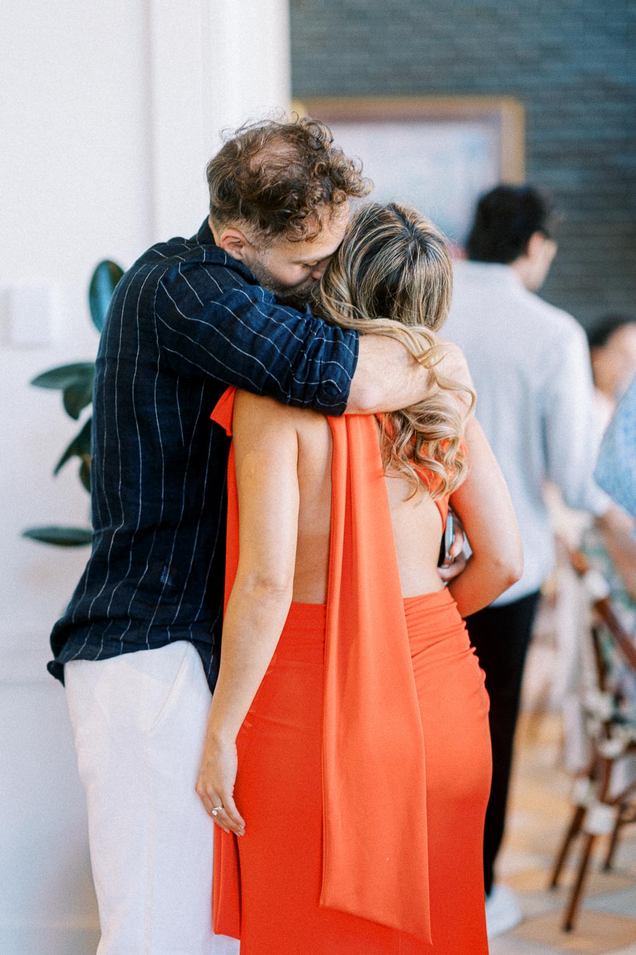 Man in striped shirt embracing woman in orange dress at a social gathering.