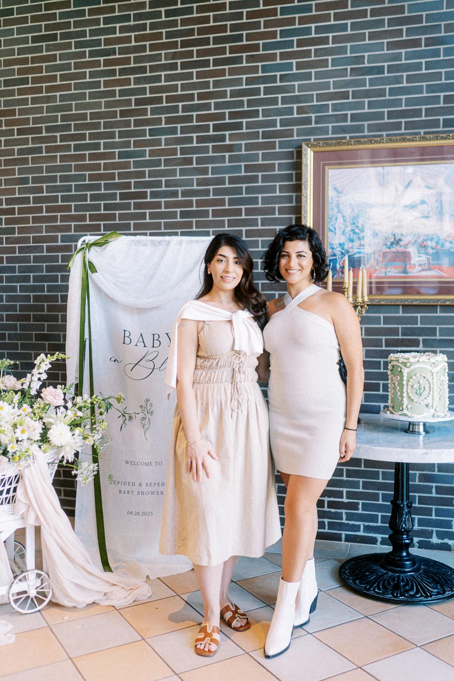 Two women posing at a stylish baby shower, featuring elegant decorations with flowers, a banner reading Baby in Bloom, and a beautifully decorated cake on a stand.