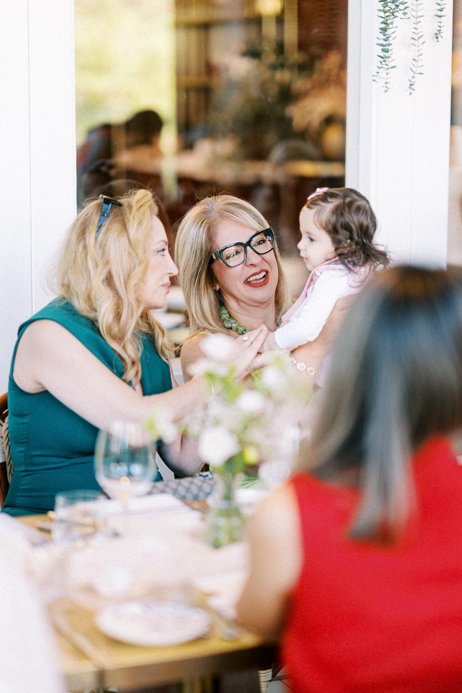 Two women smiling and interacting with a baby at a dining table set for a social gathering, with floral decorations in the foreground.