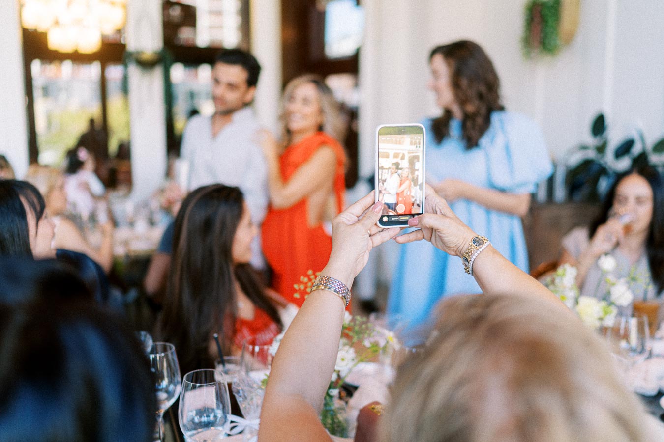 A group of people at a lively social gathering, with a focus on a person taking a photo using a smartphone. The scene captures a warm and festive atmosphere with guests interacting around a beautifully set table with floral arrangements.