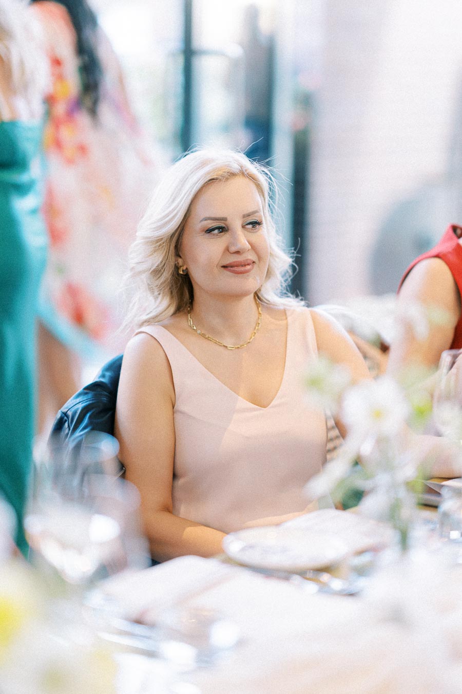 A woman with blonde hair in a pink dress sits at a dining table adorned with plates and glasses, surrounded by a softly blurred background of other guests at an elegant gathering.