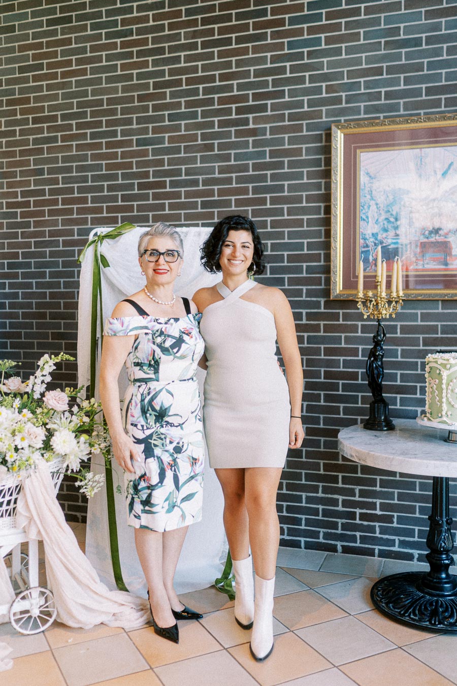 Two women posing together at a celebration event, both wearing stylish dresses. The woman on the left is older, wearing glasses and a floral dress, while the younger woman on the right wears a white dress and boots. They're standing in a decorated venue with flowers and a cake, against a brick wall background.