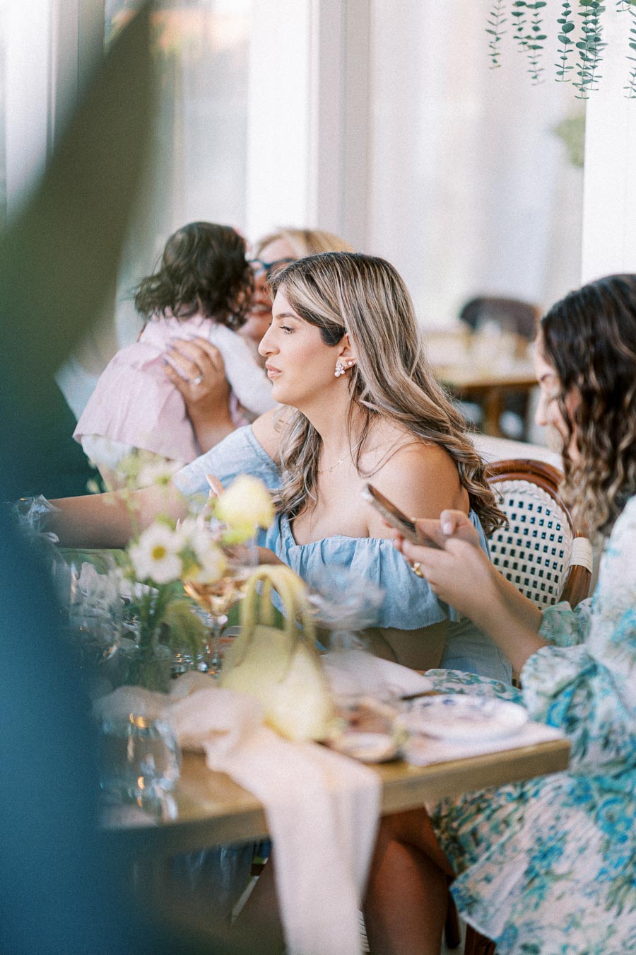 A group of people enjoying a casual gathering at a restaurant, with a woman in a blue off-shoulder top focused on her phone, surrounded by elegantly set tables and flowers, creating a warm and inviting atmosphere.