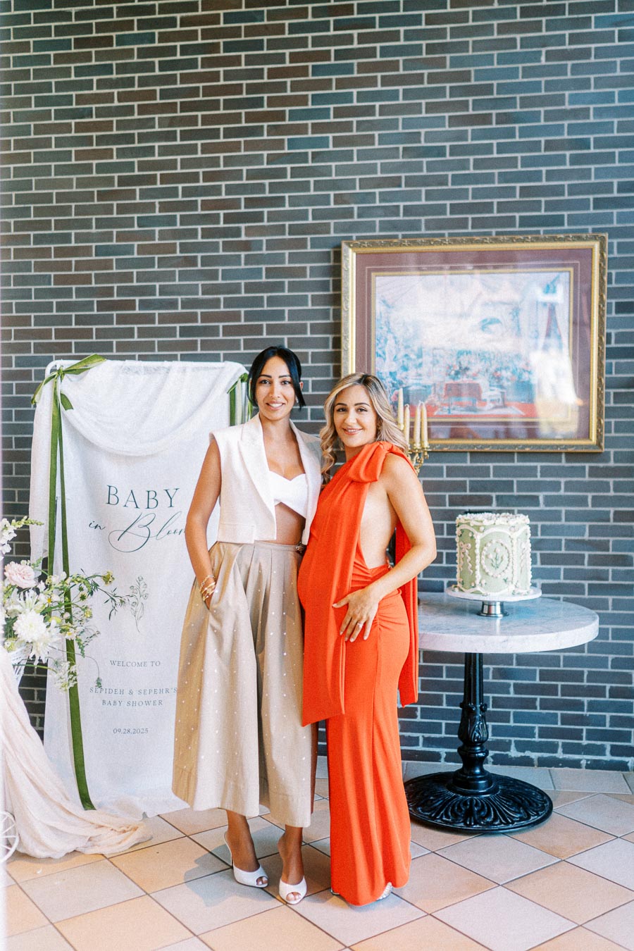 Two women standing in front of a Baby in Bloom sign at a baby shower event. One is wearing a beige skirt and white top, while the other is in a vibrant orange dress. A decorative cake is placed on a table beside them, with floral accents complementing the celebration theme.