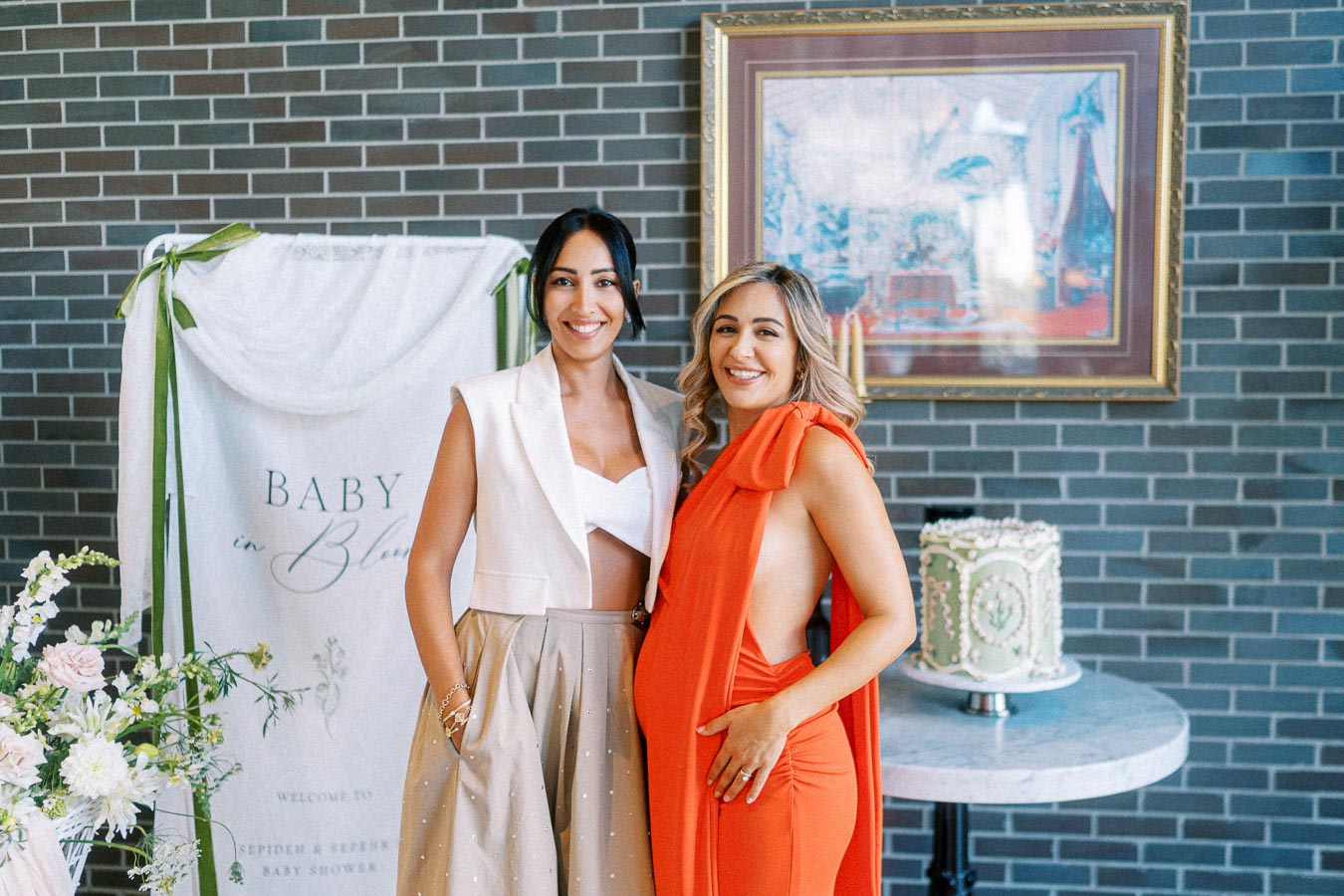 Two smiling women pose at a “Baby in Bloom” themed baby shower, with a decorative cake and floral arrangements in the background.