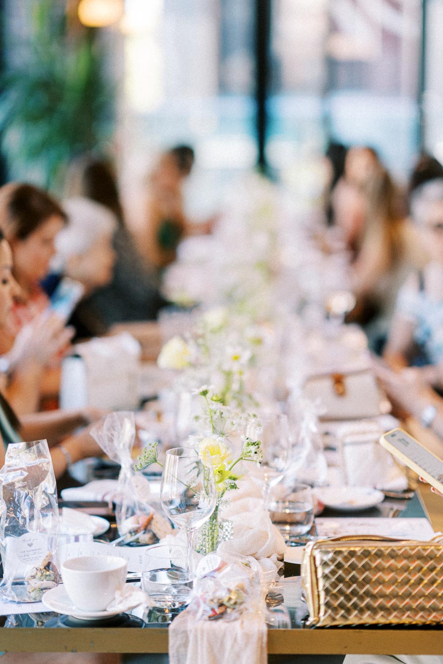A beautifully set long dining table for a social gathering, adorned with white floral arrangements, glassware, and wrapped gift favors, surrounded by people in a bright, airy restaurant setting.