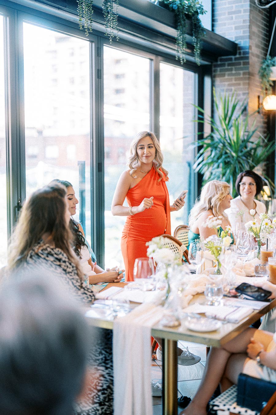 Woman in an orange dress holding a phone, standing and speaking to a group of women seated around a table during a gathering in a stylish indoor setting with large windows and greenery.