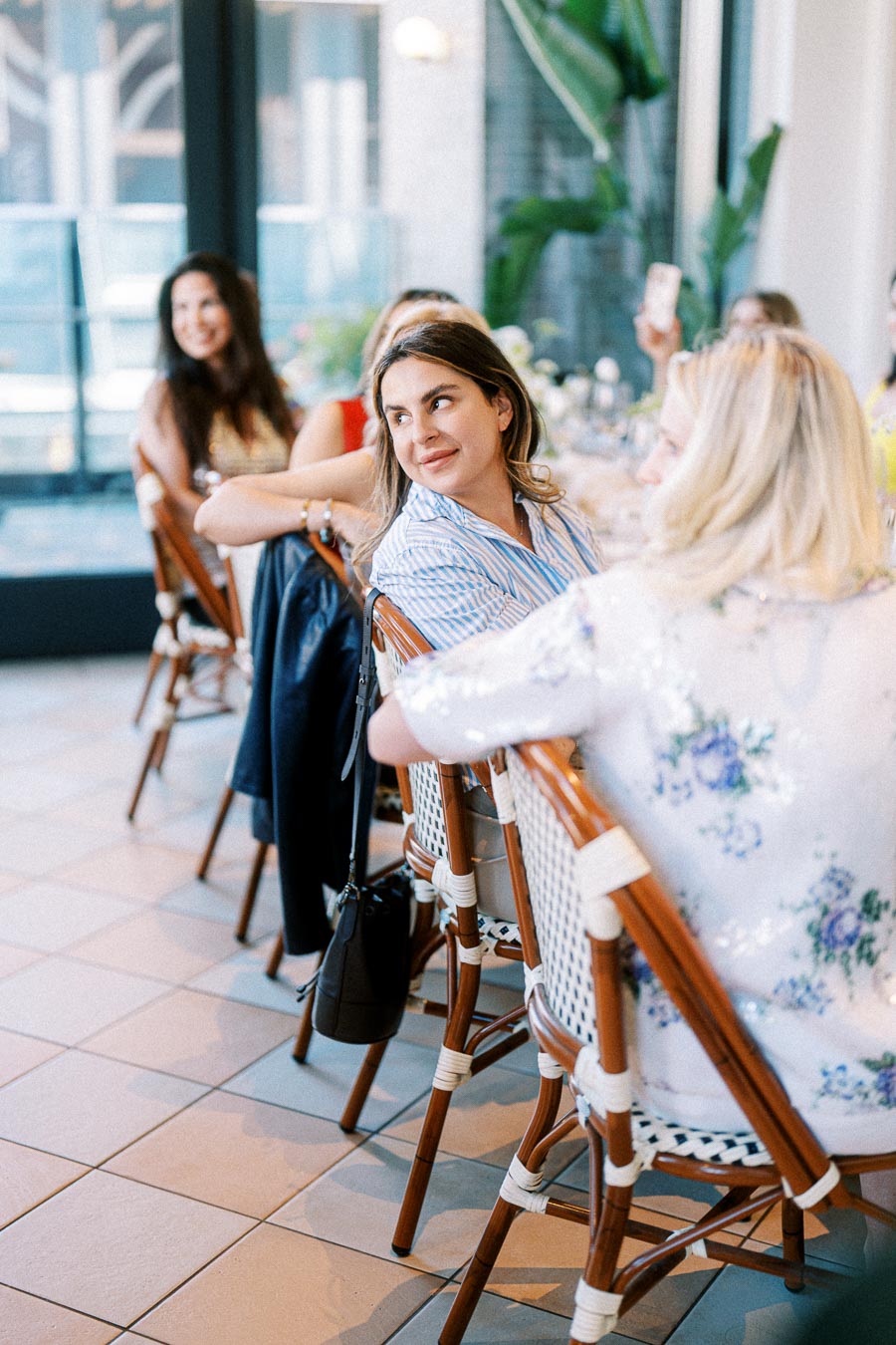 Group of women sitting on woven chairs at a stylish indoor gathering, engaging in conversation with a bright and airy setting in the background.
