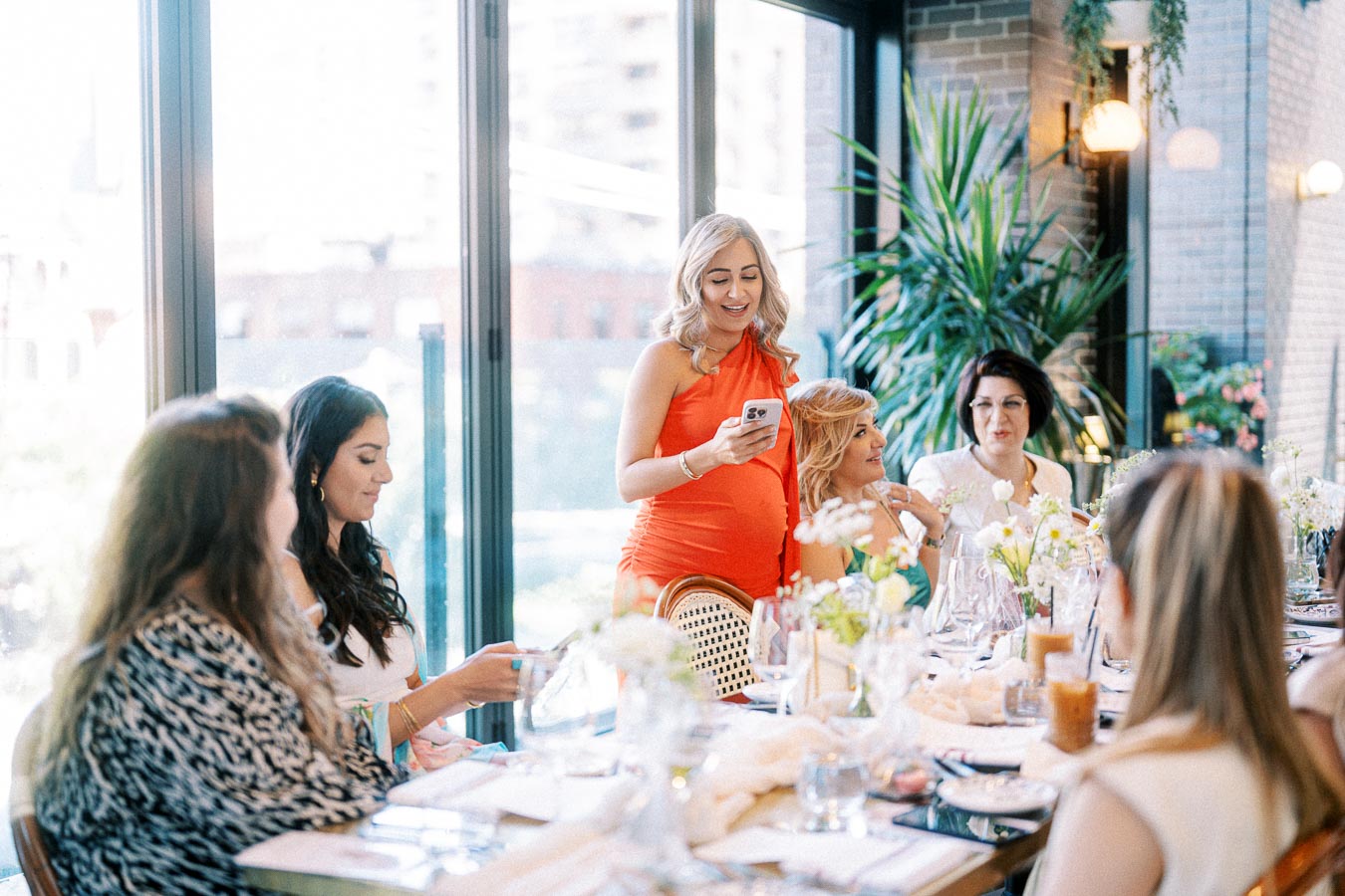A group of women enjoying a baby shower in a bright, elegant restaurant setting, with one woman standing and reading from a phone at a beautifully decorated table with flowers and glassware.