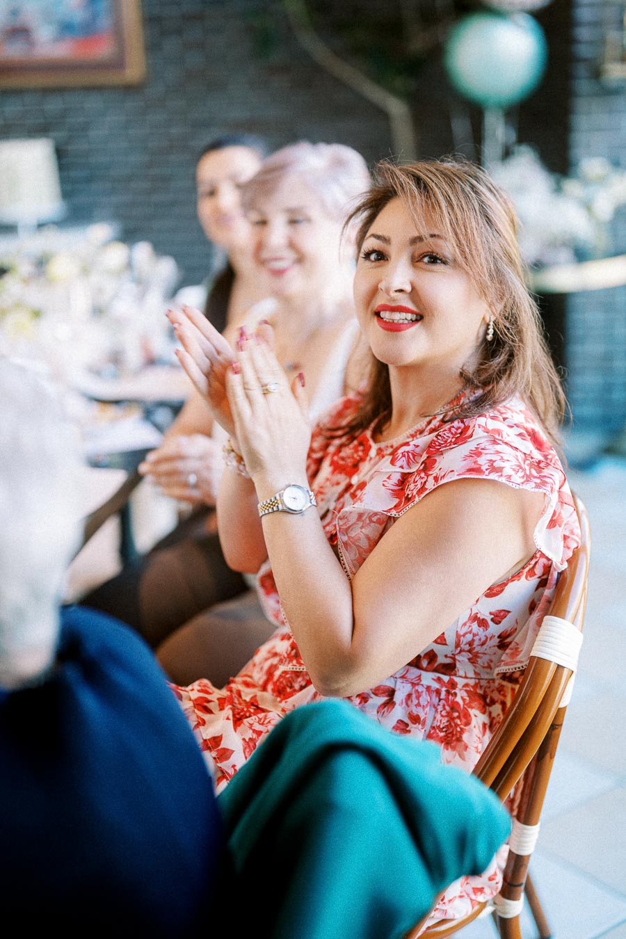 A woman in a red floral dress clapping and smiling at a joyful gathering, sitting at a decorated table with other attendees in a festive setting.