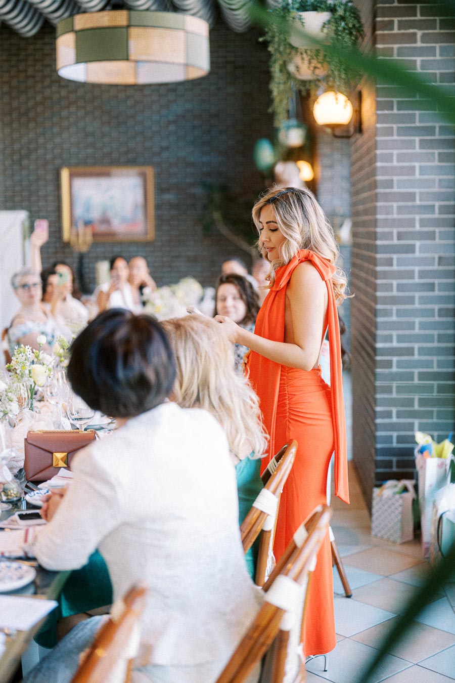 A woman in a vibrant orange dress stands giving a speech at an elegantly decorated event, surrounded by seated guests at a long table with floral arrangements and fine dining settings.