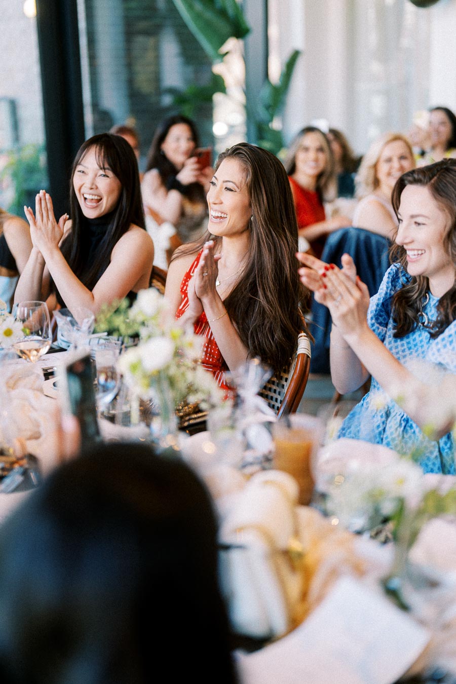 Group of people clapping and smiling at a lively gathering, seated around a beautifully decorated table with flowers and drinks.