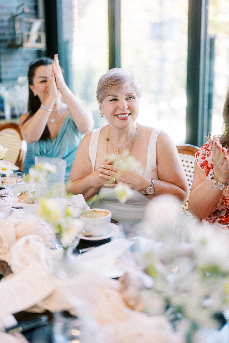 Smiling elderly woman at a lively dining table, adorned with flowers and enjoying brunch with friends in a bright, sunlit café.