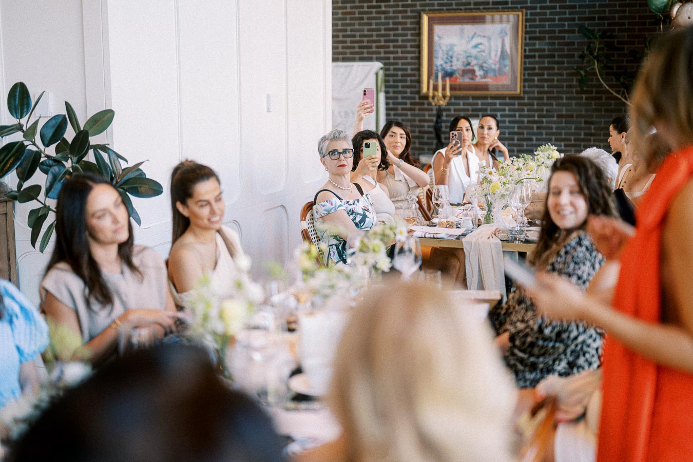 A lively bridal shower gathering with guests sitting at a beautifully decorated table, capturing moments on their phones.