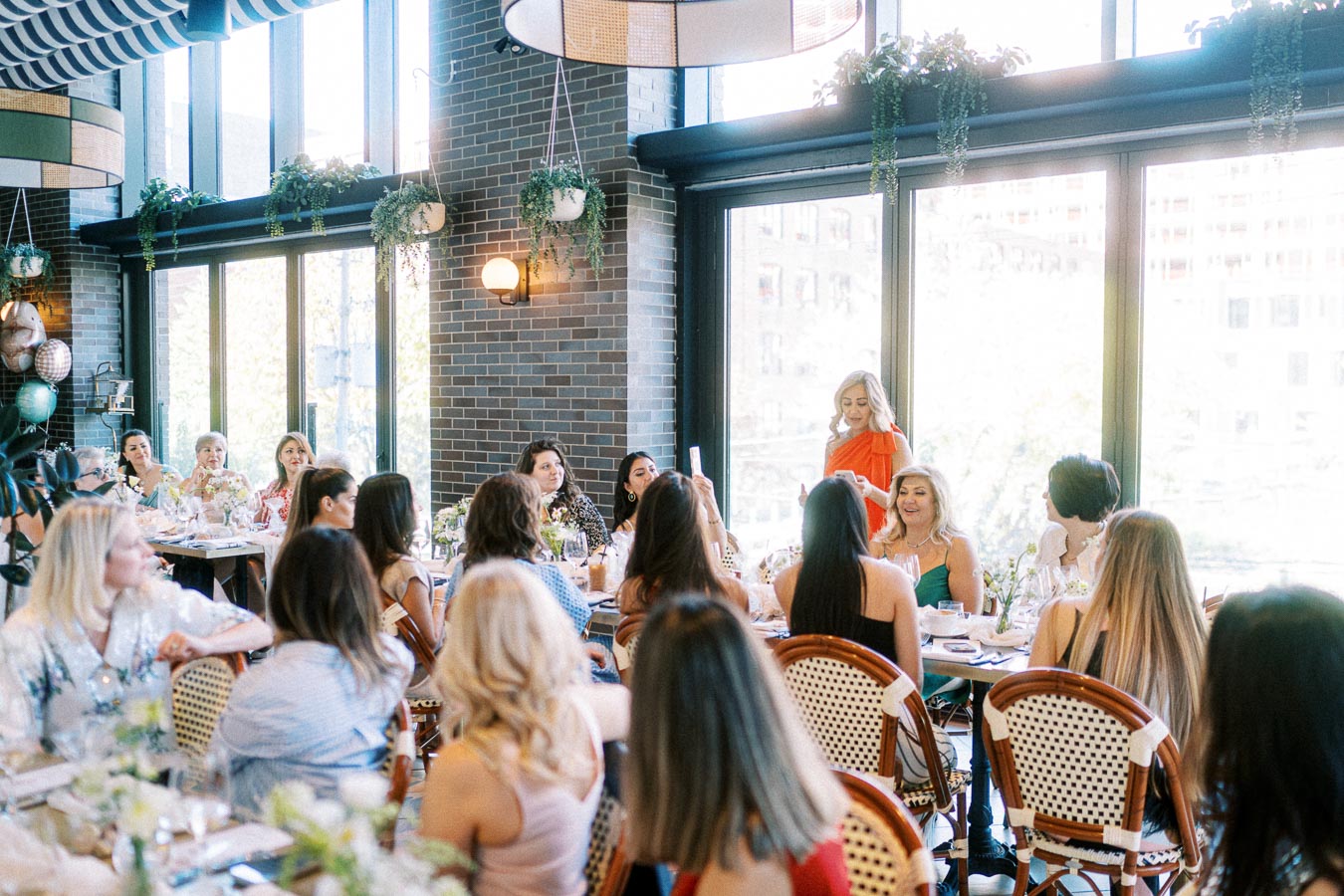 Elegant gathering of women at a stylish restaurant event, with a speaker in a bright orange dress addressing the group, surrounded by modern decor and large windows.