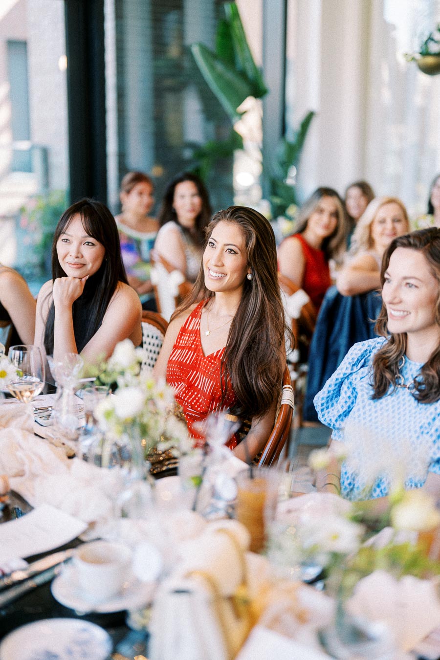 A group of women smiling and enjoying a social gathering at a beautifully set table with floral arrangements in a bright, indoor setting.