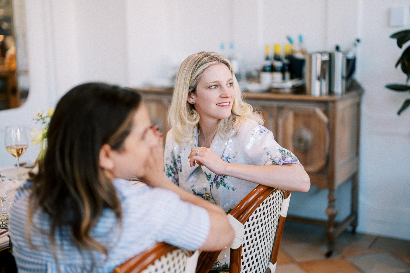 Two women sitting at a table, engaged in conversation in a cozy, elegantly decorated room with wine bottles in the background.