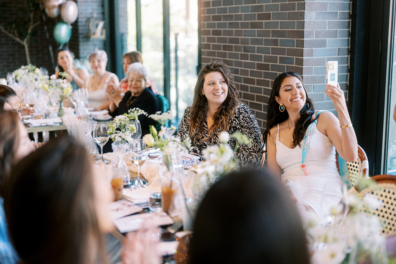 A group of women smiling and enjoying a celebration at a beautifully decorated table with flowers and drinks, one taking a selfie, suggesting a joyful social gathering in a bright, modern setting.