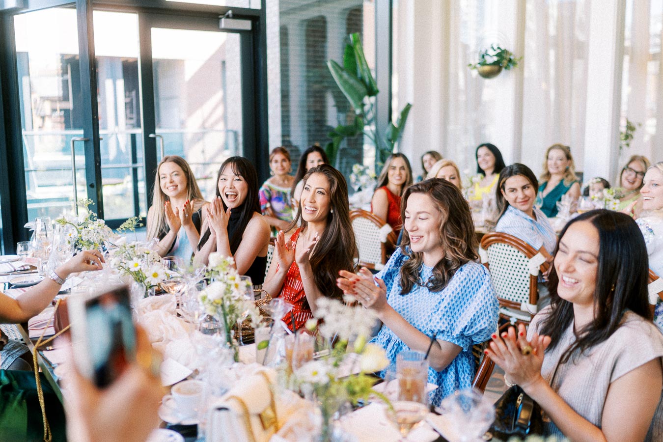Group of smiling people enjoying a celebration at a beautifully set table in a bright, modern venue with flowers and glassware.