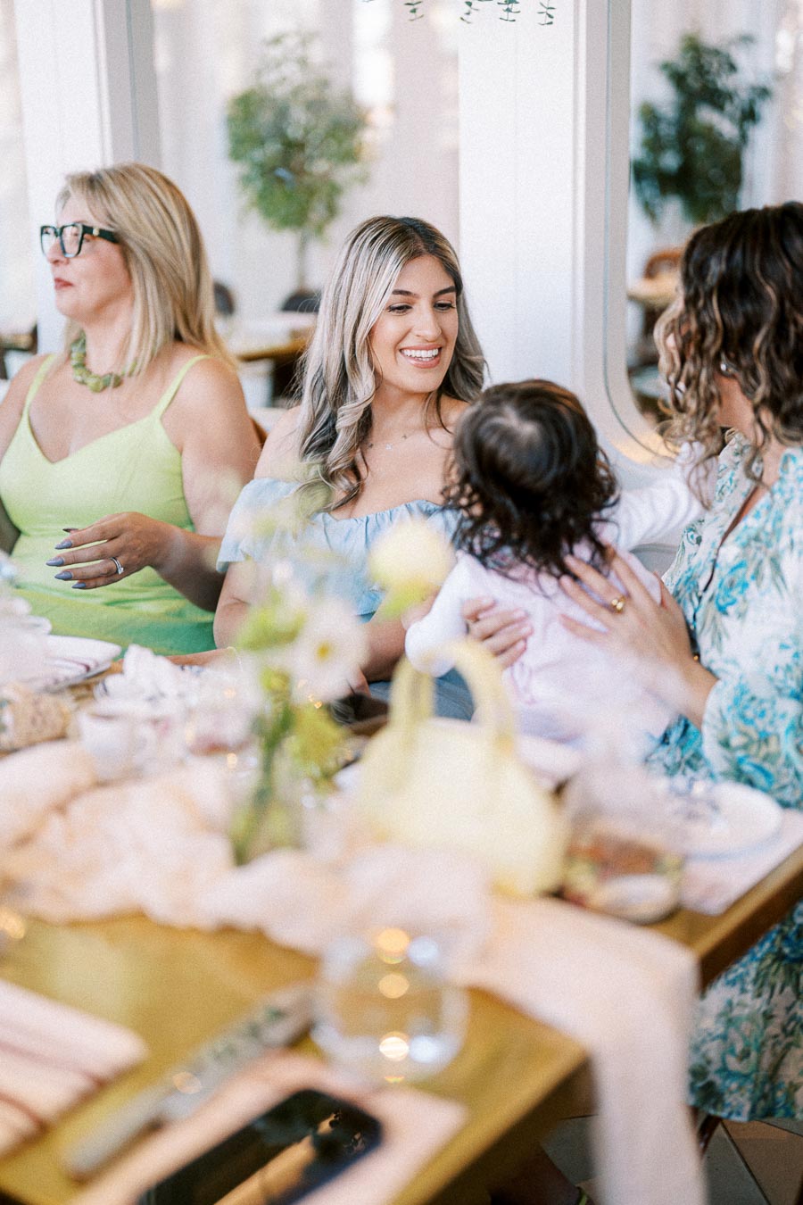 A group of women enjoying a cheerful brunch with a toddler at a beautifully set table, surrounded by flowers and decor in a bright restaurant.