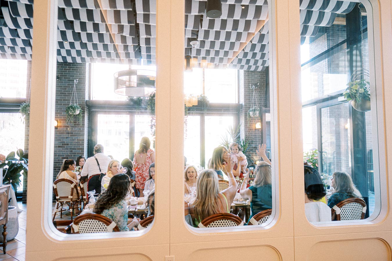 A lively gathering in a stylish modern cafe with people socializing around a table, featuring large glass windows, patterned ceiling, and greenery accents for a relaxed ambiance.